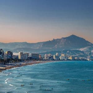 Una vista aerea di una spiaggia affollata lungo una città costiera, con una grande catena montuosa sullo sfondo sotto un cielo limpido.
