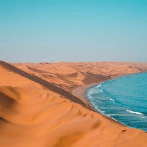 Vaste dune di sabbia arancione incontrano l'oceano blu lungo una costa sotto un cielo sereno.