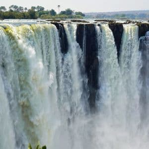 Una cascata ampia e potente precipita da un dirupo roccioso, generando una grande nuvola di nebbia alla sua base con alberi in lontananza.