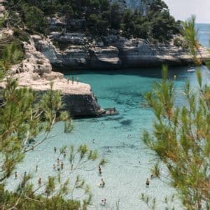 Vue aérienne de baigneurs dans une crique turquoise et transparente, bordée de falaises rocheuses et de verdure luxuriante.