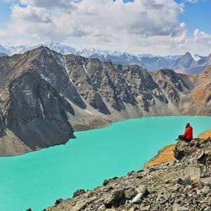 Une personne en veste rouge assise sur une falaise rocheuse surplombant un lac alpin turquoise vif entouré de montagnes sous un ciel nuageux.