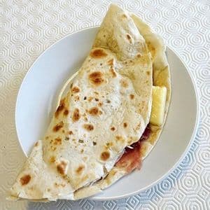 Una piadina tostata, ripiegata e ripiena di prosciutto e formaggio, su un piatto bianco.