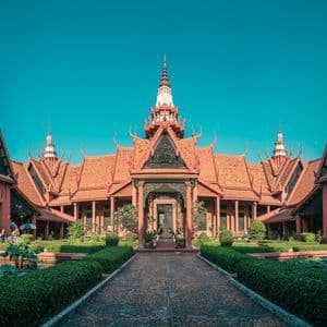 A terracotta-colored building with traditional ornate roofs stands behind a formal garden with a central stone path under a clear blue sky.