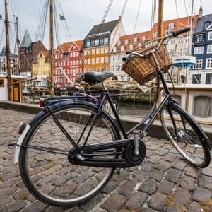 Un vélo noir avec un panier en osier est garé dans une rue pavée le long d'un canal avec des bateaux et des bâtiments colorés.