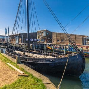 Une réplique de drakkar noir amarrée dans un port, à côté d'une jetée en bois et de bâtiments, par une journée ensoleillée.