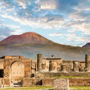 Antiche rovine con colonne e archi in pietra si ergono di fronte a un grande vulcano sotto un cielo parzialmente nuvoloso.
