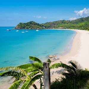 A high-angle view of a tropical bay with a white sand beach, clear turquoise water, and sailboats anchored offshore.