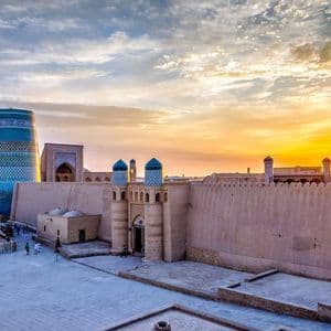 Una ciudad antigua con altas murallas de fortaleza y un gran minarete de azulejos azules brilla durante una puesta de sol dorada.