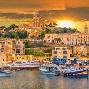 A harbor filled with boats and yachts in front of a hillside town featuring a large church under a golden sunset.