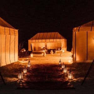 Lanterns and torches illuminate a path between large canvas tents at a desert camp during the night.