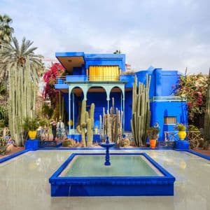 Un vibrante edificio azul se alza en un jardín de altos cactus y palmeras, con un estanque reflectante y una fuente al frente.