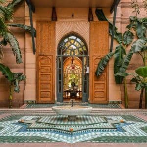 A central star-shaped fountain with green and blue mosaic tiles in a courtyard with lush plants and an arched doorway.