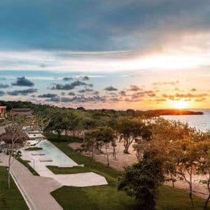 An aerial view of a coastal resort with modern buildings, pools, and trees overlooking the ocean at sunset.