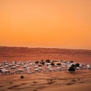 Un camp de tentes blanc se dresse dans une enceinte clôturée, au milieu de dunes de sable ondulantes, sous un ciel orangé.