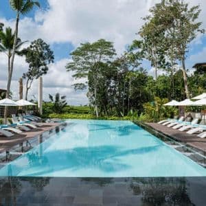 Une longue piscine dans un complexe tropical, flanquée de rangées de chaises longues blanches et de parasols sous un ciel partiellement nuageux.