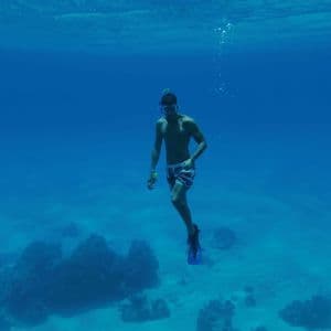 Un hombre con máscara y aletas de buceo está sumergido en aguas cristalinas y azules sobre un fondo marino arenoso con rocas.