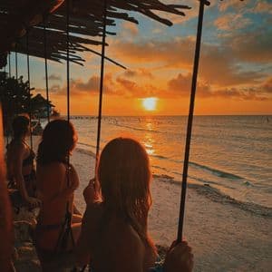 Des voyageurs d'un groupe WeRoad sont assis sur des balançoires sous un toit de chaume, contemplant le coucher de soleil sur l'océan.