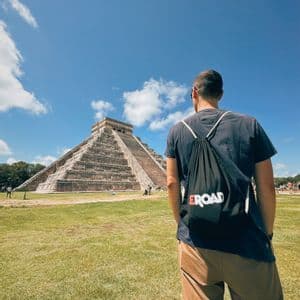 Un homme vu de dos avec un sac à dos WeRoad, contemplant une grande pyramide à degrés en pierre sur un champ herbeux sous un ciel bleu.