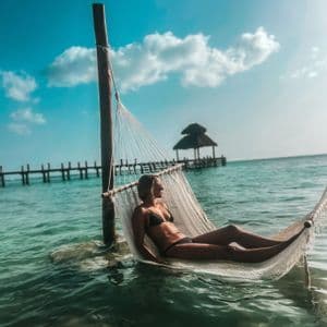 Une femme en bikini noir se détend dans un hamac blanc suspendu au-dessus de l'eau turquoise de l'océan avec une jetée en arrière-plan.