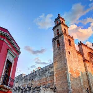 Una vista en contrapicado de una gran catedral de piedra iluminada por el atardecer, junto a un vibrante edificio rojo bajo un cielo parcialmente nublado.