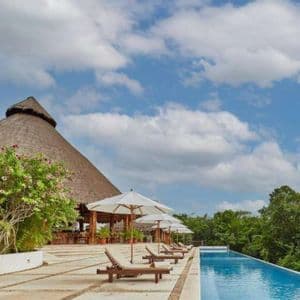 Sonnenliegen mit Sonnenschirmen säumen einen Infinity-Pool in einem Resort, dessen reetgedecktes Gebäude einen Wald überblickt.