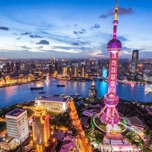 Una vista aerea di una torre futuristica illuminata di rosa che domina uno skyline tentacolare e un ampio fiume al crepuscolo.