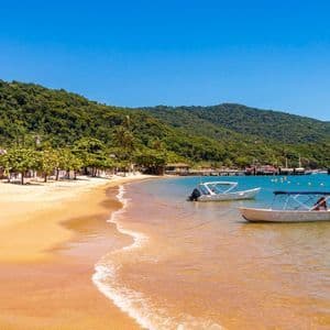 Due piccole barche sono ancorate nelle acque limpide di una baia tranquilla, accanto a una spiaggia sabbiosa alberata e con alle spalle una grande collina boscosa.