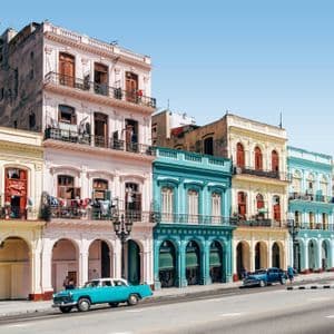 Un'auto d'epoca blu brillante percorre una strada fiancheggiata da colorati edifici coloniali a più piani sotto un cielo sereno.