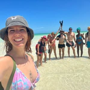 Eine lächelnde Frau mit Fischerhut macht ein Selfie mit einer WeRoad-Gruppenreise im seichten, klaren türkisfarbenen Wasser stehend.