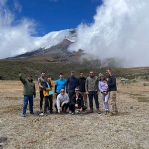 A WeRoad group trip poses for a photo in a mountain landscape with a large, snow-capped peak partially covered by clouds.