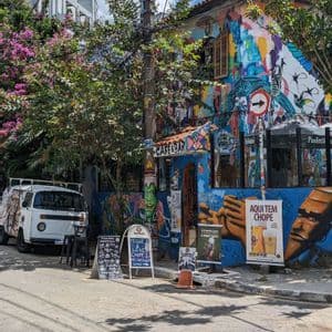 Un café y bar de esquina en una calle de la ciudad, sus paredes cubiertas de coloridos murales y grafitis, con una furgoneta blanca aparcada cerca.