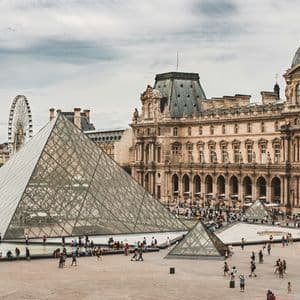Folla di persone si raduna in un grande cortile attorno a una grande piramide di vetro, con un edificio storico sullo sfondo.