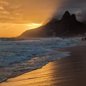 Ozeanwellen schlagen bei Sonnenuntergang an einem Sandstrand auf, wobei goldenes Licht auf dem Wasser reflektiert wird und sich dahinter silhouettierte Berge abzeichnen.