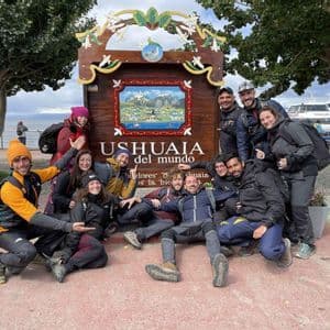 Eine WeRoad-Gruppe posiert lächelnd am Ufer vor dem großen Holzschild 'Ushuaia del mundo'.