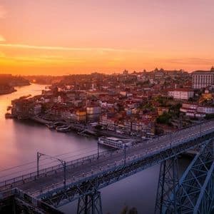 Un grande ponte ad arco metallico attraversa un ampio fiume, accanto a un paesaggio urbano sotto un cielo vivace al tramonto arancione.