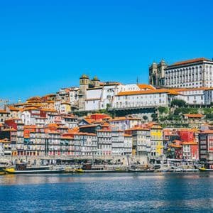 Una città colorata in collina con tetti arancioni è visibile da oltre un fiume sotto un cielo azzurro limpido.