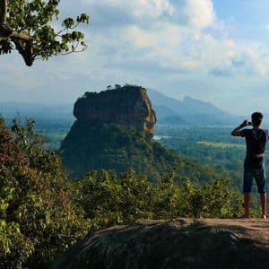 Da un punto di vista elevato, una persona fotografa un'imponente formazione rocciosa circondata da una fitta foresta verde e montagne.