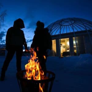Due persone di un viaggio di gruppo WeRoad si stagliano contro un falò nella neve al tramonto, con un igloo di vetro sullo sfondo.