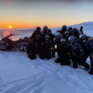 Un gruppo WeRoad in posa per una foto in un paesaggio innevato accanto a una motoslitta al tramonto.