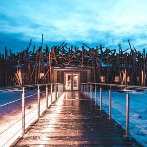 Una passerella in legno illuminata conduce a un edificio moderno con un'unica facciata a doghe, immerso in un paesaggio innevato al tramonto.