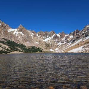 Un limpido lago di montagna riflette una catena montuosa frastagliata e innevata sotto un cielo azzurro senza nuvole.