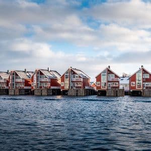 Una fila di case rosse di legno su palafitte con tetti innevati si trova sul bordo dell'acqua sotto un cielo nuvoloso.