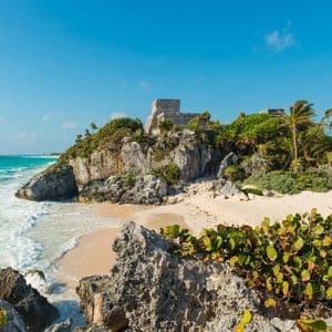 Un'antica rovina in pietra sorge su una scogliera verde a picco su una spiaggia sabbiosa, con un'iguana che riposa su una roccia vicino al mare turchese.