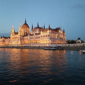 Ein prunkvolles, beleuchtetes Parlamentsgebäude an einem Flussufer in der Abenddämmerung, dessen Lichter sich im Wasser spiegeln.
