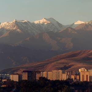Eine Stadt-Skyline mit Gebäuden liegt am Fuße sanfter Hügel, im Hintergrund eine große, schneebedeckte Bergkette.