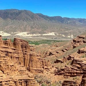 A view into a canyon with layered red rock formations, with distant dry mountains under a bright blue sky.
