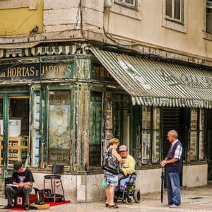 Gente su un marciapiede di città, davanti a una bottega d'angolo rustica e vissuta, con una tenda a strisce verdi e bianche.