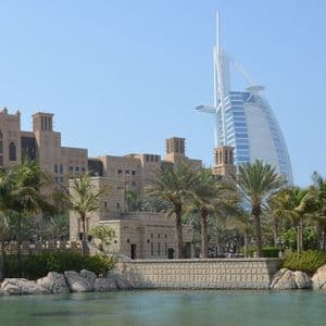 Un grattacielo a forma di vela si erge dietro edifici tradizionali color sabbia e palme, con uno specchio d'acqua calmo in primo piano.