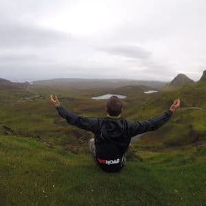 Une personne portant un sac à dos WeRoad est assise sur une colline verdoyante, les bras ouverts, contemplant une vallée parsemée de lacs sous un ciel nuageux.