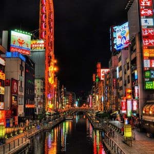 Un canale cittadino di notte fiancheggiato da edifici con insegne al neon giapponesi colorate che si riflettono sull'acqua.